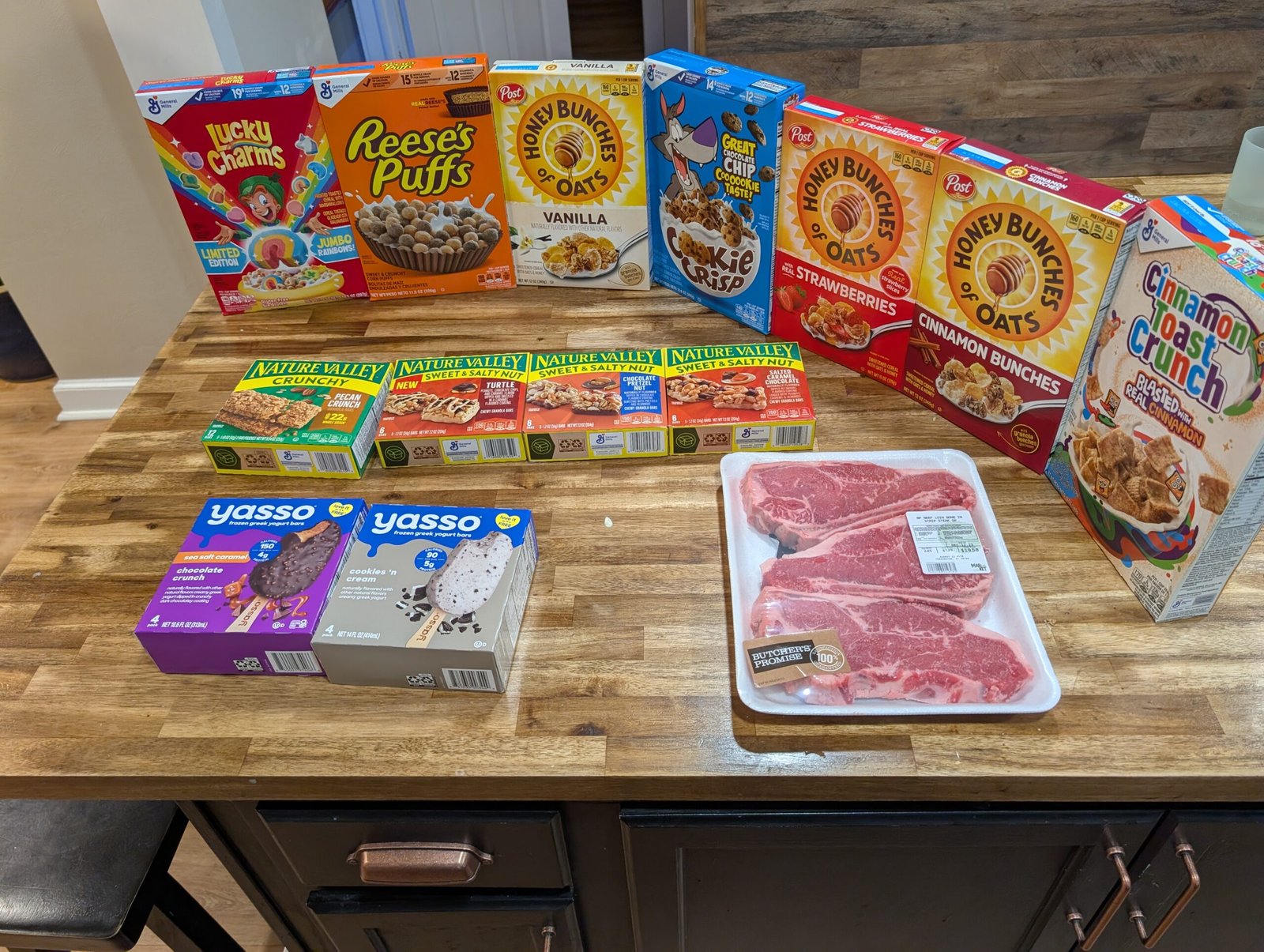 Grocery haul featuring cereals, snacks, steaks, and yogurt on the kitchen island
